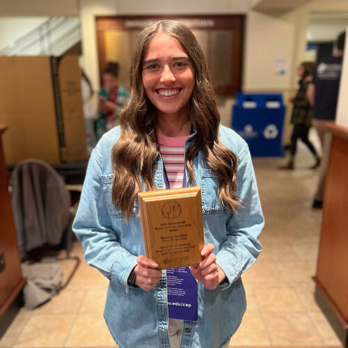 Female Teacher standing in an open exhibit space, holding a small wooden plaque with her name and school and "Sprout teacher award" etched on it.
